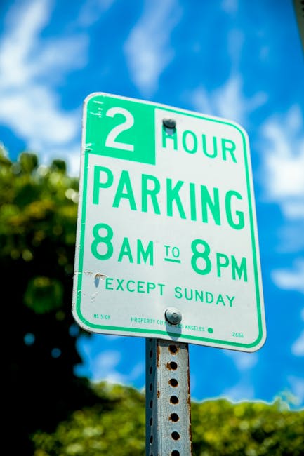 A close-up image of a green and white parking sign positioned on a metal pole against a blue sky with scattered clouds. The sign indicates a two-hour parking limit with a time restriction from 8 am to