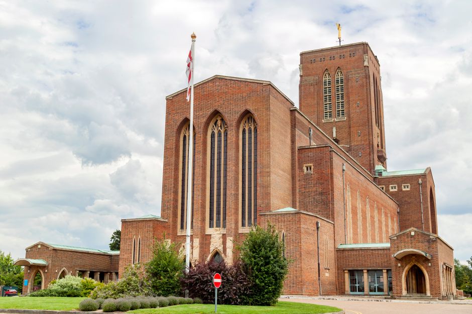 Exterior view of a large red brick church with tall, narrow Gothic-style windows and a prominent tower under a partly cloudy sky. The church is surrounded by well-maintained landscaping, including green bushes and small trees. An upright British flag on a tall pole is positioned near the building's front, with a 'no entry' traffic sign visible on the pavement. In the foreground, there are several cardboard boxes, wrapped furniture items, and moving blankets placed on the pavement, indicating a home relocation or furniture transport process. The scene is naturally lit, capturing the environment during daylight, and subtly reflects moving logistics, such as packing and loading, associated with house removals, with Surrey Movers' services aligned to house removals in Surrey, especially in areas with narrow streets like Guildford.