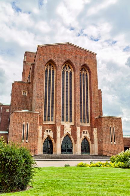 A large, red-brick church building with tall, narrow, arched stained glass windows dominating the front facade. The structure features multiple levels with small side windows and decorative stone detailing around the entrances. The building is set against a partly cloudy sky, with some sunlight illuminating the red brickwork. In front of the church, there is a well-maintained green lawn with low bushes and yellow flowers lining the edge of the grass. The scene suggests a peaceful, traditional setting, typical of architecture seen during home relocation and moving processes involving large buildings or churches, with the image capturing the exterior of a notable local landmark often included in Surrey house removals guides.