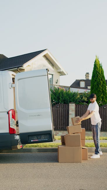 Two women are engaged in a home relocation process, sitting inside the open rear door of a large moving van with the loading area visible behind them. One woman, with long brown hair and wearing a bla